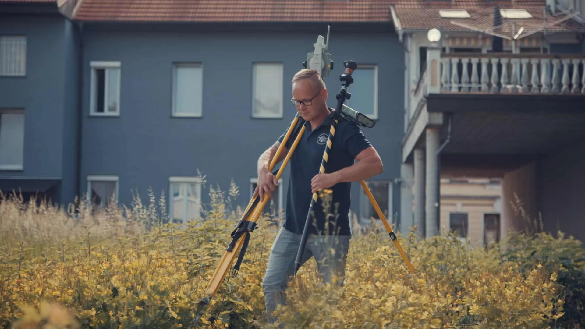 Vermessungstechniker geht durchs hohe Gras und hält ein Stativ in der Hand.