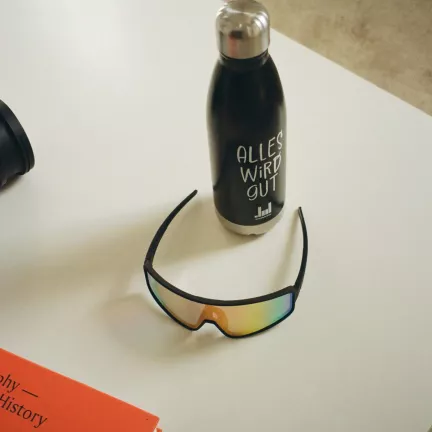 analoges foto von einer schwarzen trinkflasche mit der aufschrift "alles wird gut" und einer sportbrille auf einem weißen tisch